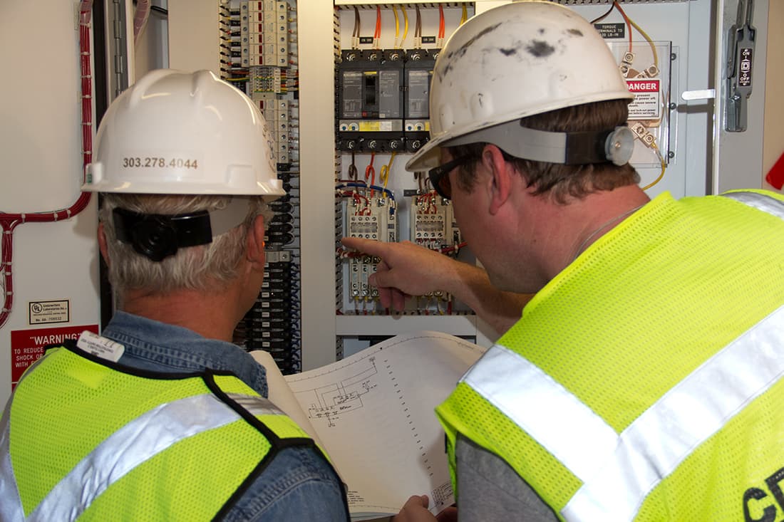 Two Karsten electricians in hard hats and hi-vis vests reading a drawing at an open control panel. The phone number 303.278.4044 is printed on one hard hat.
